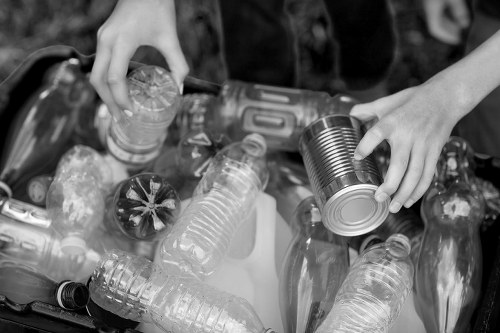 A pair of hands is shown handling a metal can, placing it among a collection of various discarded plastic bottles and containers, which are scattered across the surface. The plastic bottles are transparent, featuring different shapes and sizes, with some still having caps attached while others are open. The bottles include standard water and fizzy drink bottles, with textured surfaces and ridges typical of such containers. The scene appears outdoors, possibly on a pavement or driveway, with the background slightly blurred. The overall context suggests waste collection or sorting, aligning with rubbish removal services offered by House Clearance Reading in the Reading area. Lighting is natural, providing clear visibility of the bottles' textures and the metallic sheen of the can. The image emphasizes the importance of proper waste management and recycling practices within the local context of Reading, Berkshire.