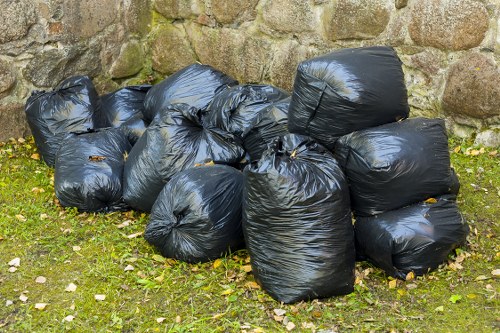A collection of ten black trash bags made of thick plastic material, tied securely at the top, situated outdoors on a grassy area with small yellow and brown leaves scattered around. The bags are arranged in two overlapping rows, resting on the ground close to a weathered stone wall composed of irregularly shaped stones in shades of brown, beige, and grey. The background shows the stone wall extending upwards, with some moss growth visible on the stones. The scene appears to be part of a rubbish removal or clearance process, typical of services provided by House Clearance Reading in the Reading area, with natural lighting suggesting daytime conditions.