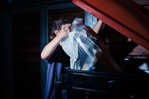A person wearing a dark shirt is seen disposing of a large clear plastic bag filled with various items into a black rubbish skip or container. The setting appears to be an outdoor area, possibly a driveway or designated waste disposal point, with a wooden structure and blue wooden panels visible in the background. The person is lifting the plastic bag towards the container’s opening, which is partially covered by a red lid that is tilted back. The scene is illuminated by natural or ambient lighting, highlighting the textures of the plastic, the person’s arms, and the surrounding environment. This image relates to waste and rubbish removal services often provided by companies like House Clearance Reading, serving areas including Reading and nearby localities, with a focus on general rubbish disposal tasks.