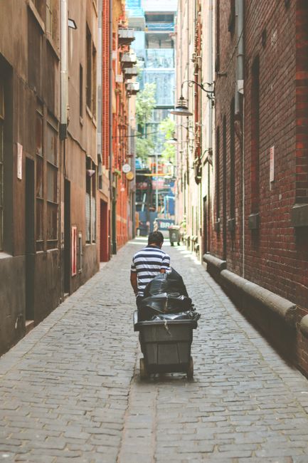 A narrow urban alleyway between two multi-storey brick buildings, with dark shadows cast by the structures on each side. The alley features a slightly sloped asphalt surface with double yellow lines r