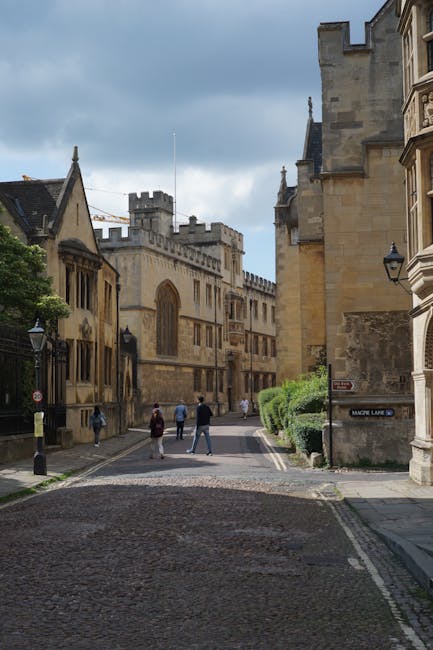 A cobblestone street scene in a historic city area featuring medieval and Renaissance-style stone buildings with ornate facades and narrow windows. The buildings are constructed from beige and light b